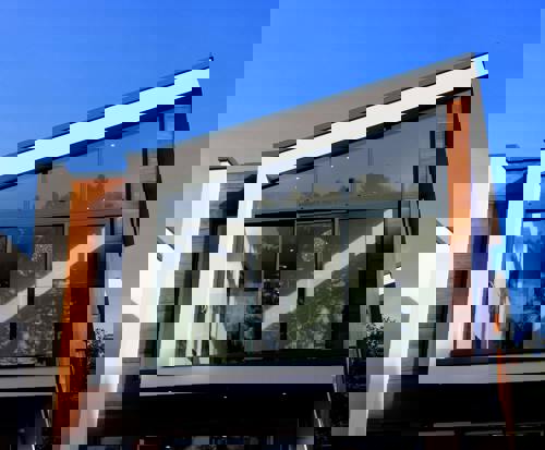 logo Modern tvåplans villa med stora glaspartier, träpanel och vit putsfasad mot blå himmel.