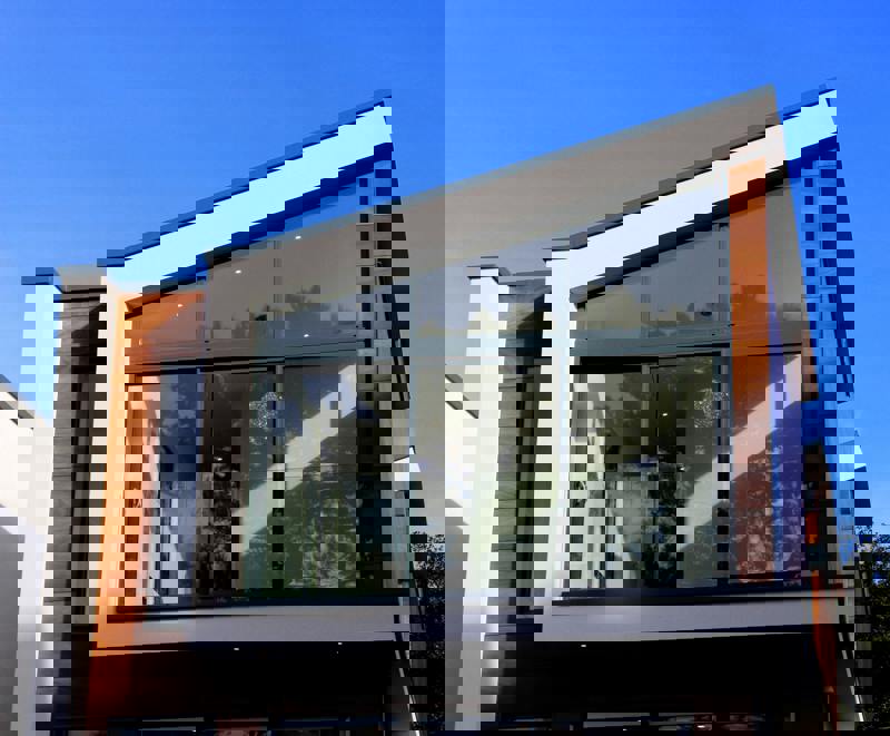 Modern tvåplans villa med stora glaspartier, träpanel och vit putsfasad mot blå himmel.