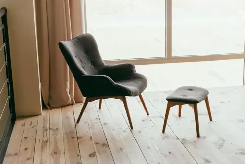 logo En elegant mörkgrå fåtölj med träben och matchande fotpall står på ett ljust trägolv, placerad vid ett stort fönster med utsikt och ljusa gardiner som ramar in rummet.