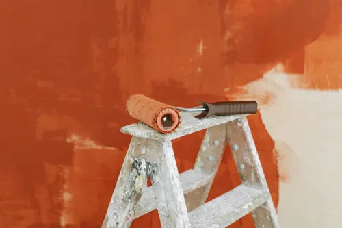 logo Målarroller på en stege/målarbock framför en vägg som målas i terrakottafärg.