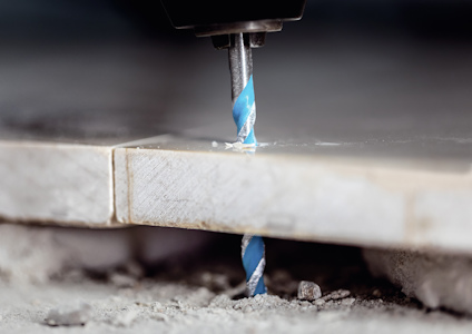 Borrmaskin borrar genom en kakelplatta, med blå spiralborr och damm runt omkring.