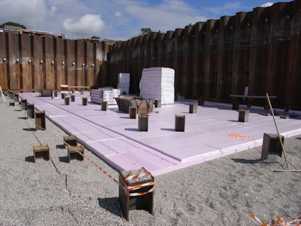 Byggarbetsplats med rosa isoleringsmaterial på marken, omgiven av höga stålspontväggar.