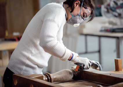 En person i vit tröja och ansiktsmask arbetar med träbearbetning vid ett arbetsbord.
