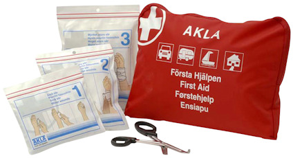 Röd första hjälpen-kudde med vit text och symboler, omgiven av förpackningar och saxar.