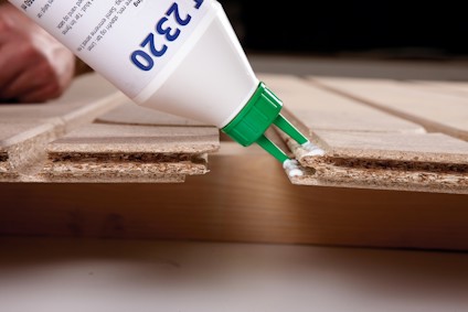 En hand applicerar lim från en tub med etiketten "P 1320" på spånskivor.