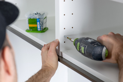 En person använder en skruvdragare för att sammanfoga vita skåpstommar med inredningsskruvar. I bakgrunden syns en öppen ask med små skruvar.