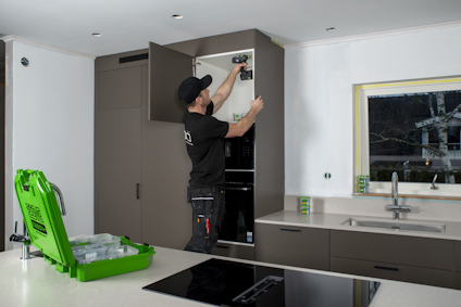 En person sammanfogar en skåpstomme med skivmaterial i ett kök. På köksön står en öppen verktygsväska med flera askar med skruvar.