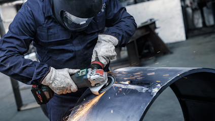 Person med skyddsutrustning slipar metall med vinkelslip, gnistor flyger.