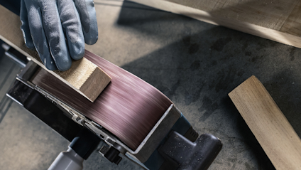Hand med skyddshandske slipar träbit mot roterande slipband.